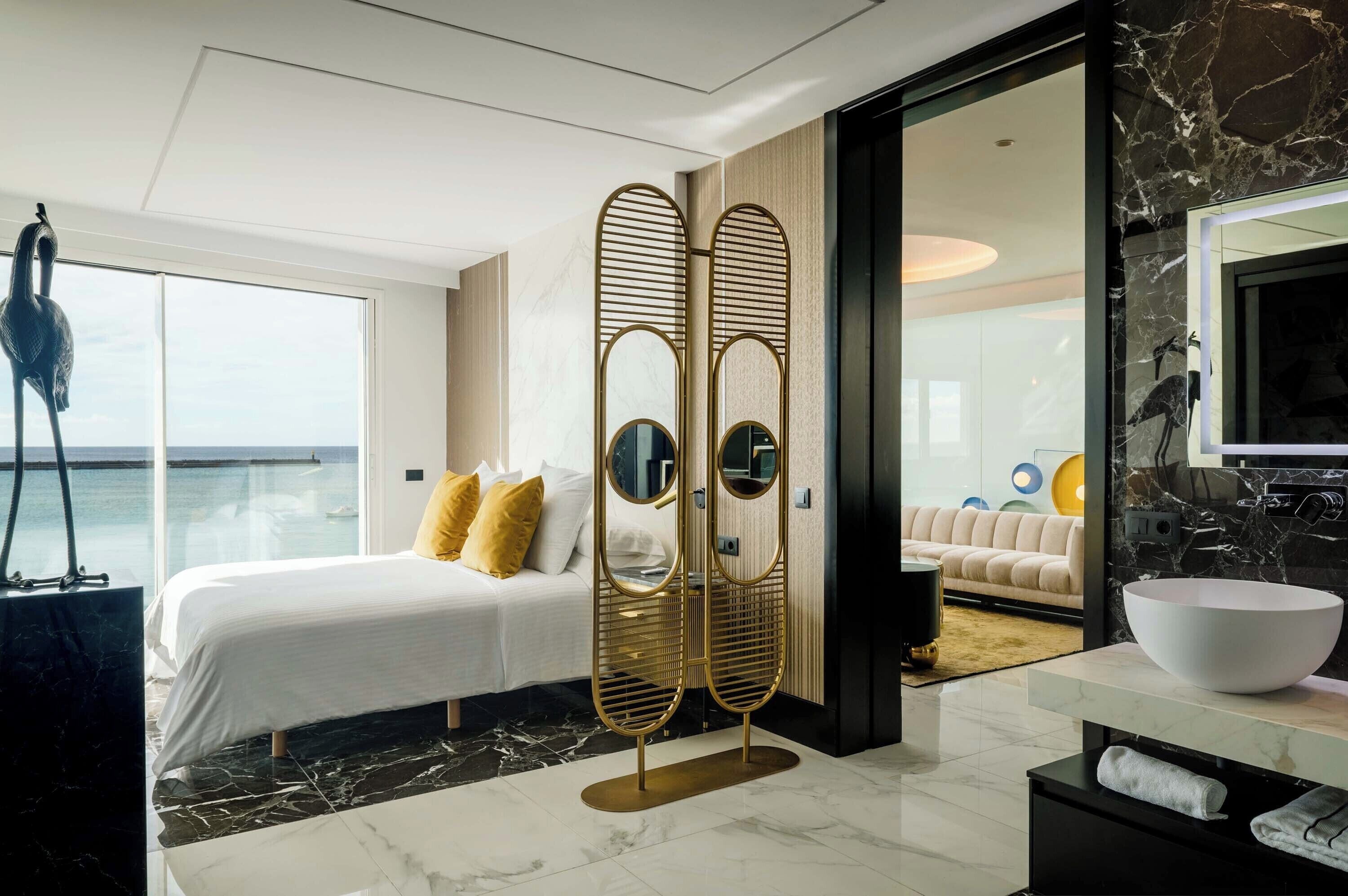 Una habitación de hotel moderna con una cama grande, vista al mar, un espejo con marco dorado, un lavabo blanco y suelo de mármol.