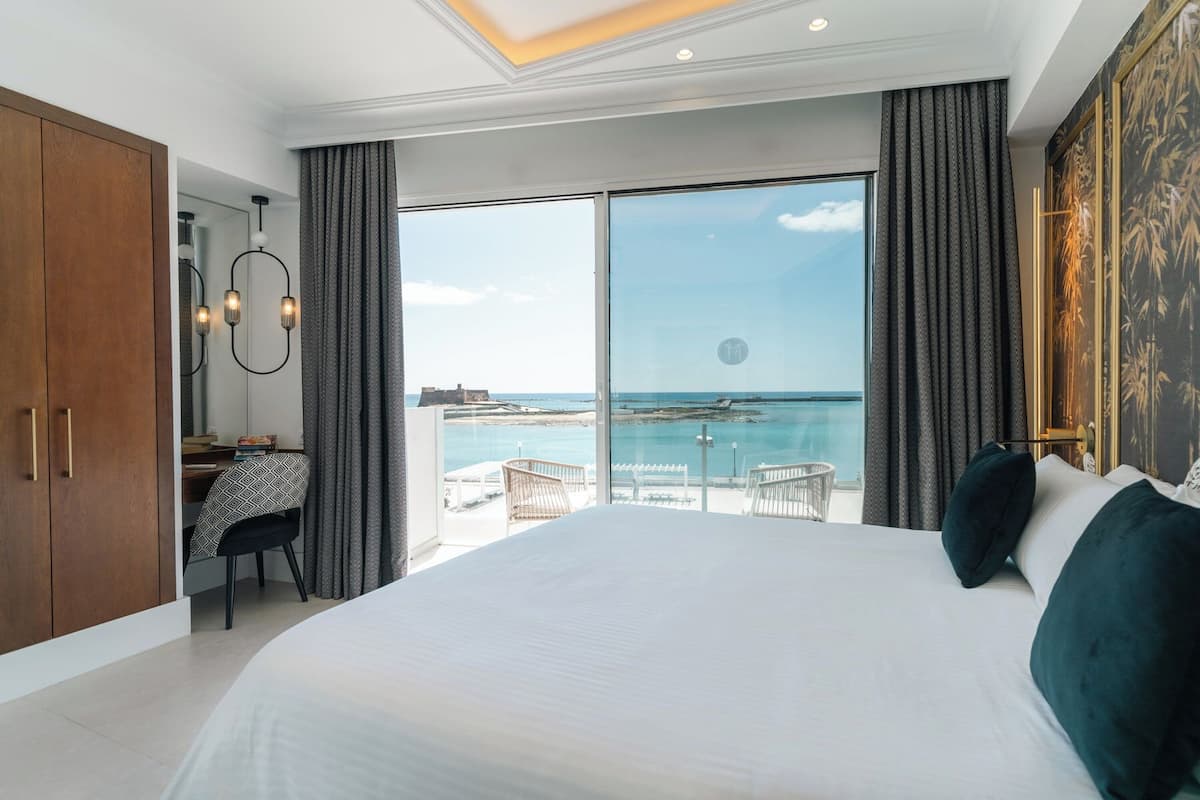A modern bedroom with a large bed, a desk, and a view of the sea.