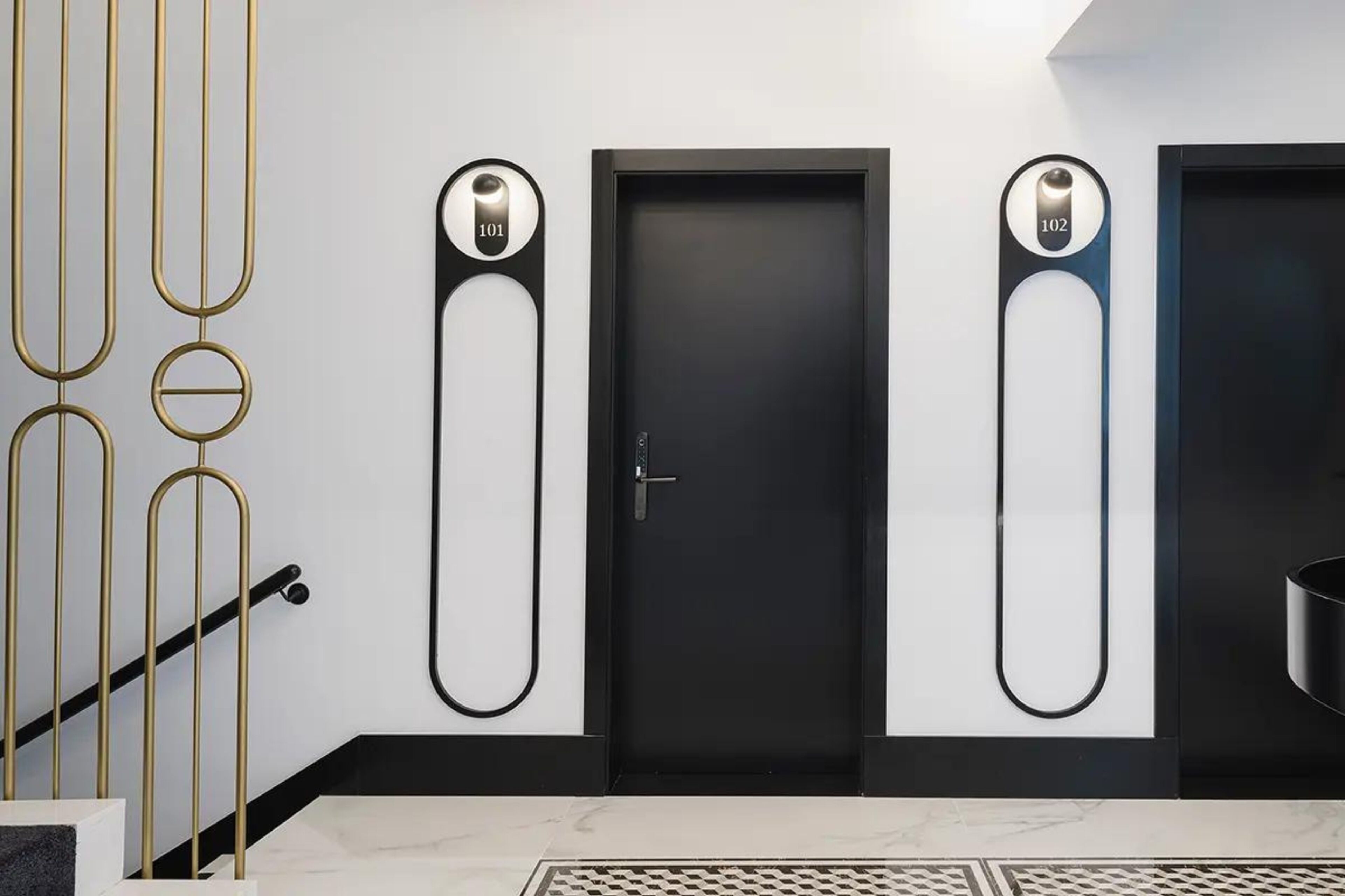 A modern hotel lobby with a black door, two elevator call buttons, and a staircase with a gold railing.