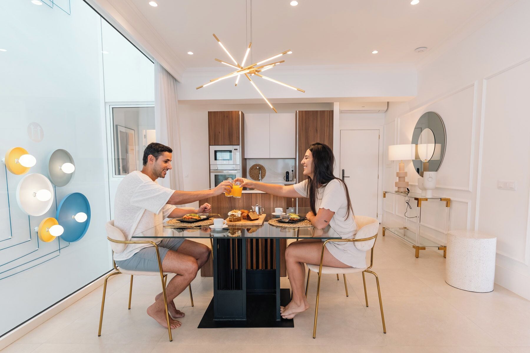 Un espace repas moderne avec une table en verre, deux personnes assises, et une cuisine en arrière-plan.