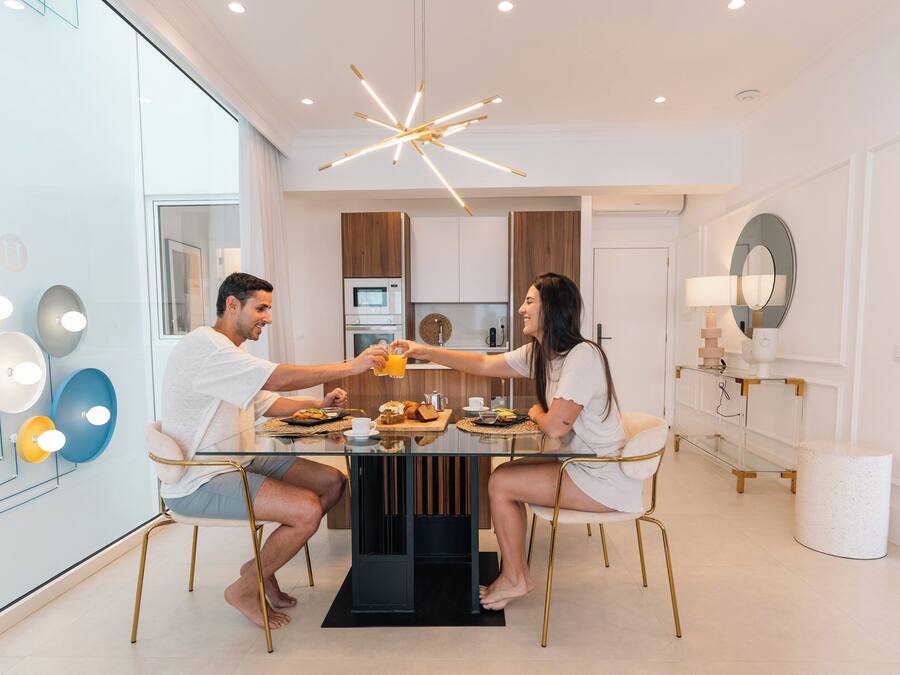 Un comedor moderno con una mesa de vidrio, dos personas sentadas y una cocina al fondo.