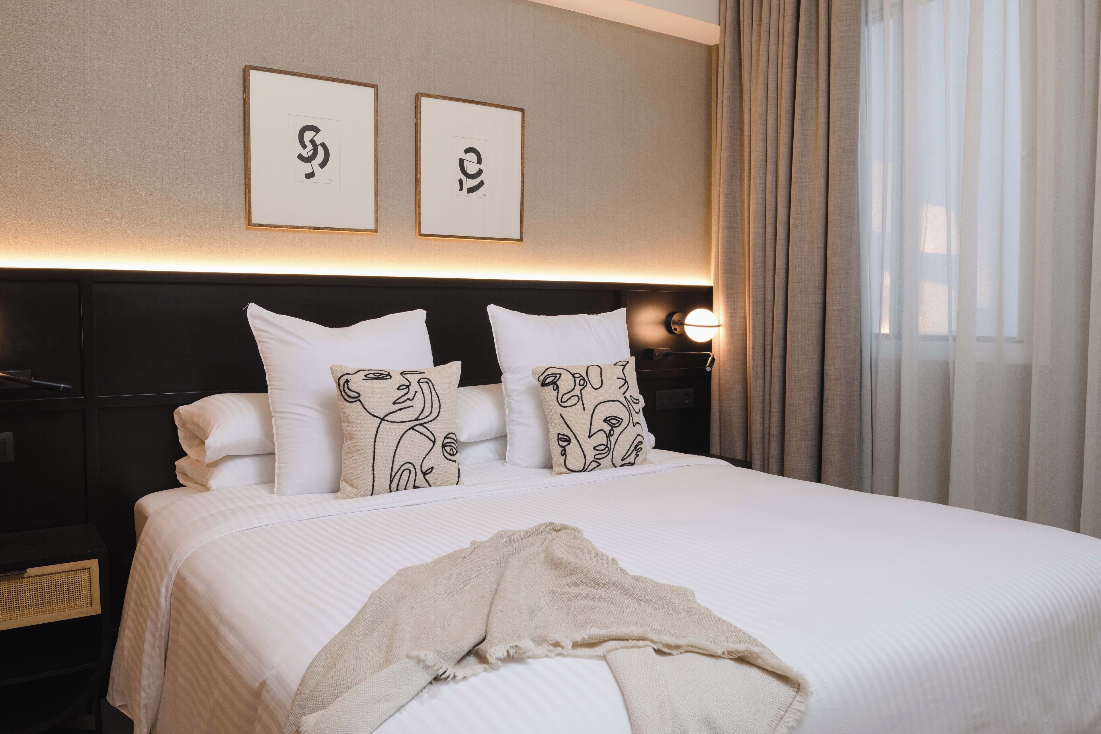 Camera d'albergo con un letto grande, due opere d'arte appese alla parete, una lampada da comodino e una finestra con tende.