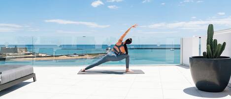 Uma pessoa fazendo uma pose de yoga em um tapete, com vista para a piscina e a praia.