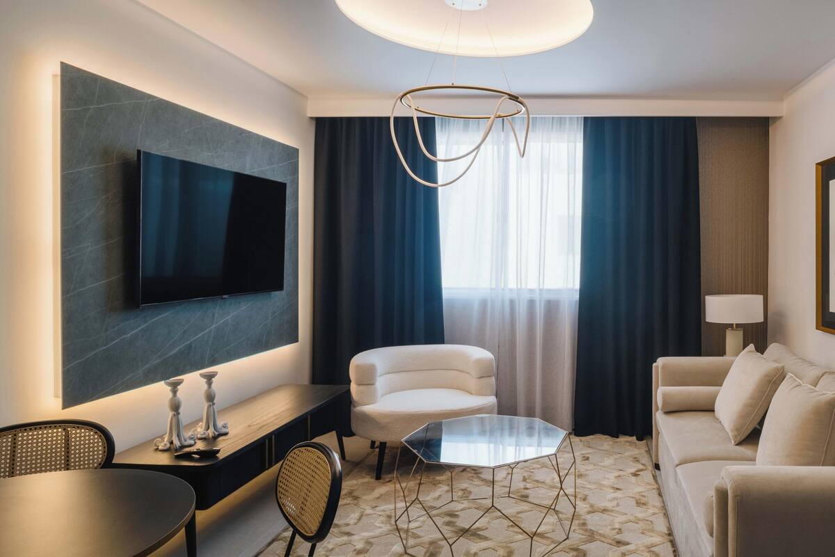 A modern hotel room with a flat-screen TV, a glass coffee table, a sofa, a chair, and a dining table.