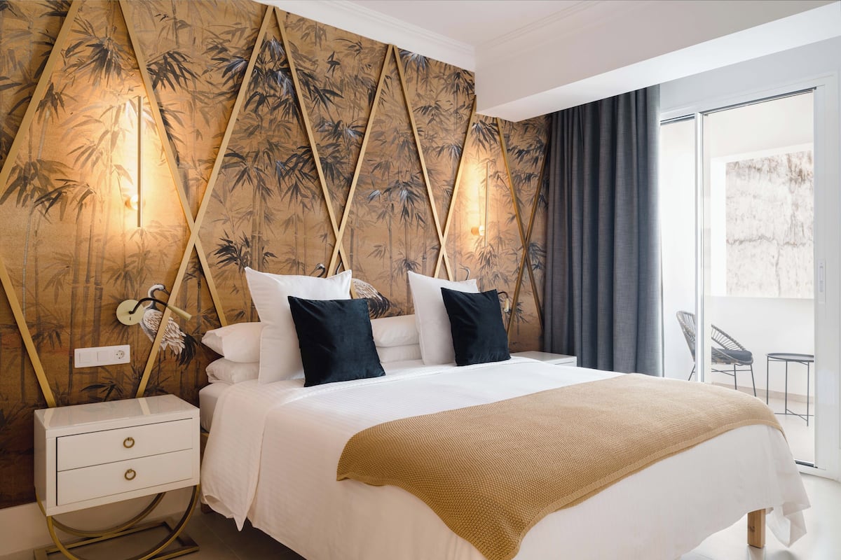 A hotel room with a large bed, a nightstand, a lamp, and a view of the outside.