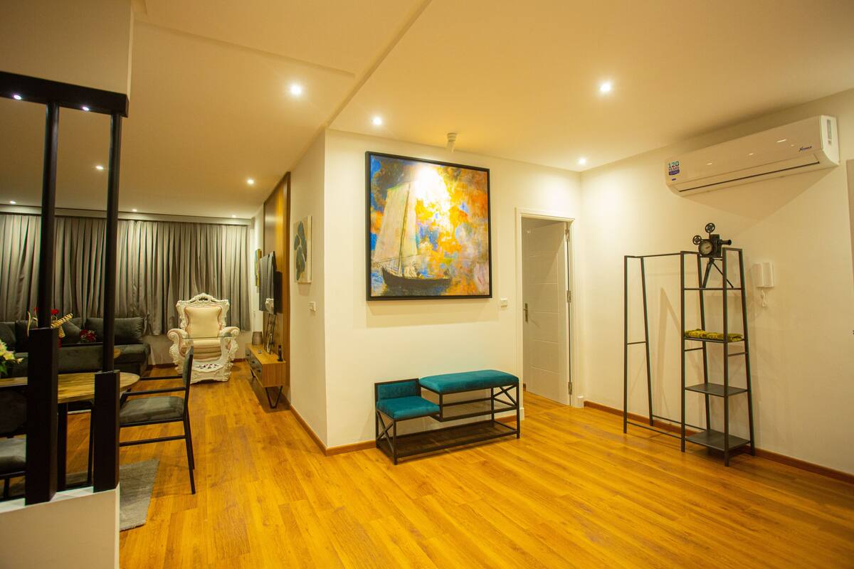 A spacious living room with a wooden floor, a large painting, a bench, a chair, and a small table.