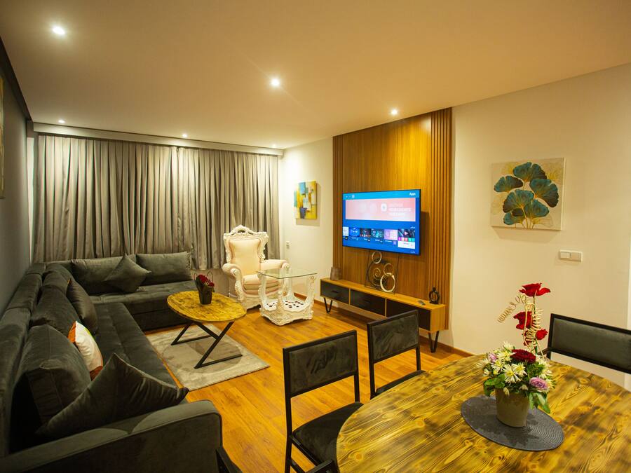 Uma sala de estar moderna com um sofá seccional, uma mesa de jantar de madeira e uma TV fixada na parede.
