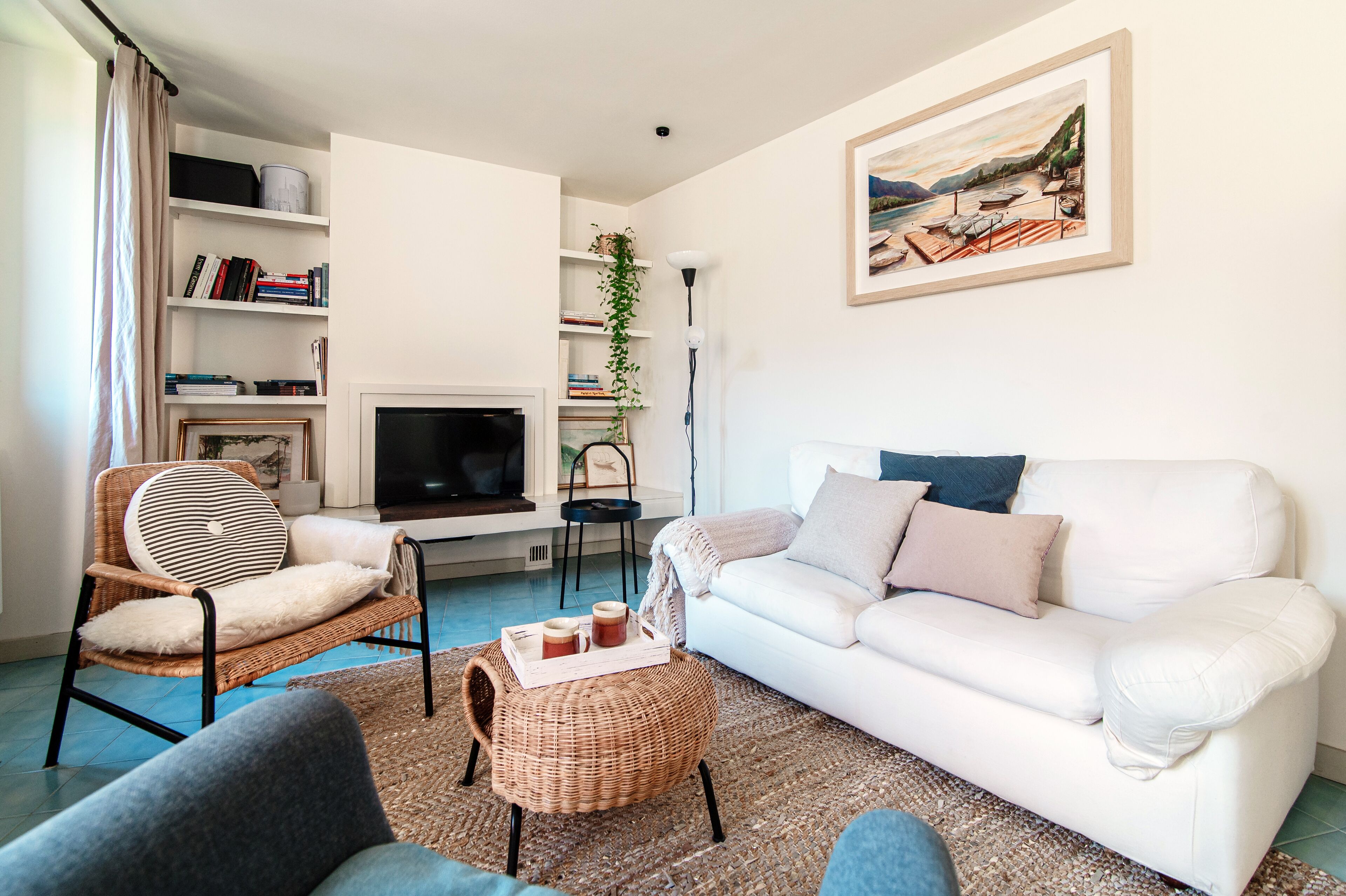 Ein Wohnzimmer mit einem weißen Sofa, Korbstühlen, einem Couchtisch mit Getränken, einem Bücherregal, einem Fernseher und einem Gemälde an der Wand.