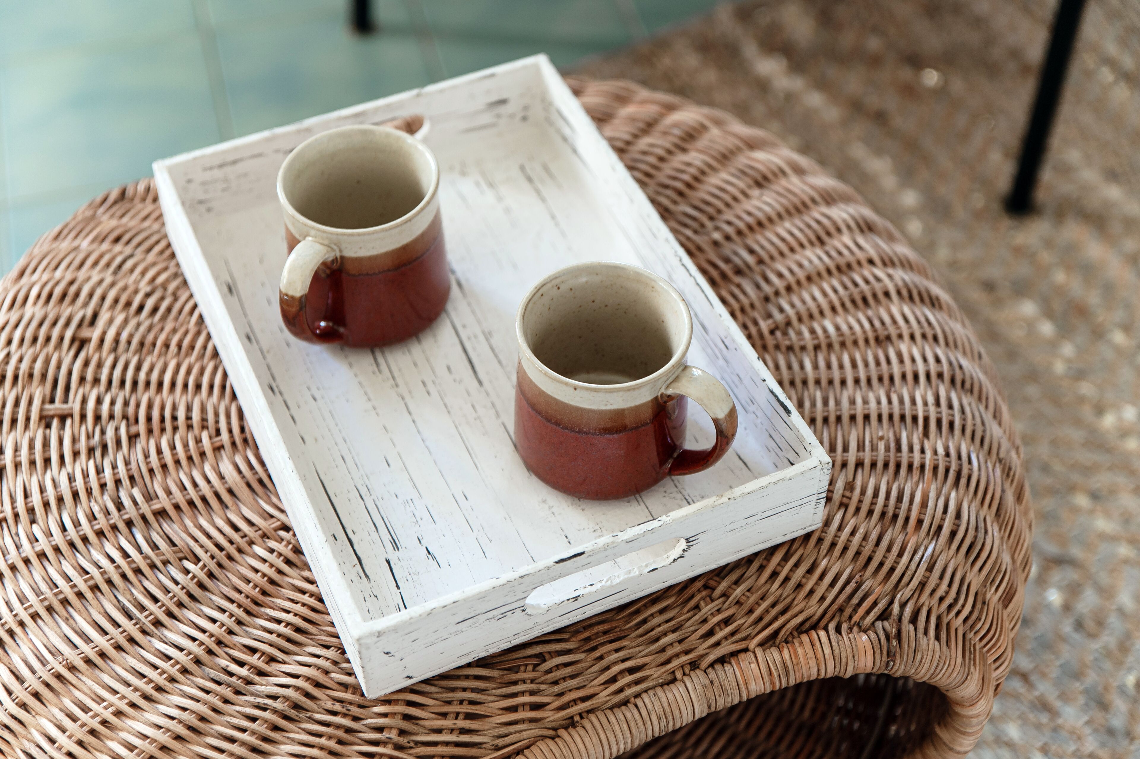 A rustic wooden tray with two ceramic mugs on a woven round stool.