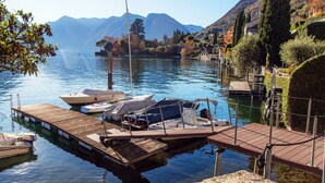 A calm lake with boats docked at a wooden pier, surrounded by hills and trees. - Olea (Tremezzina)