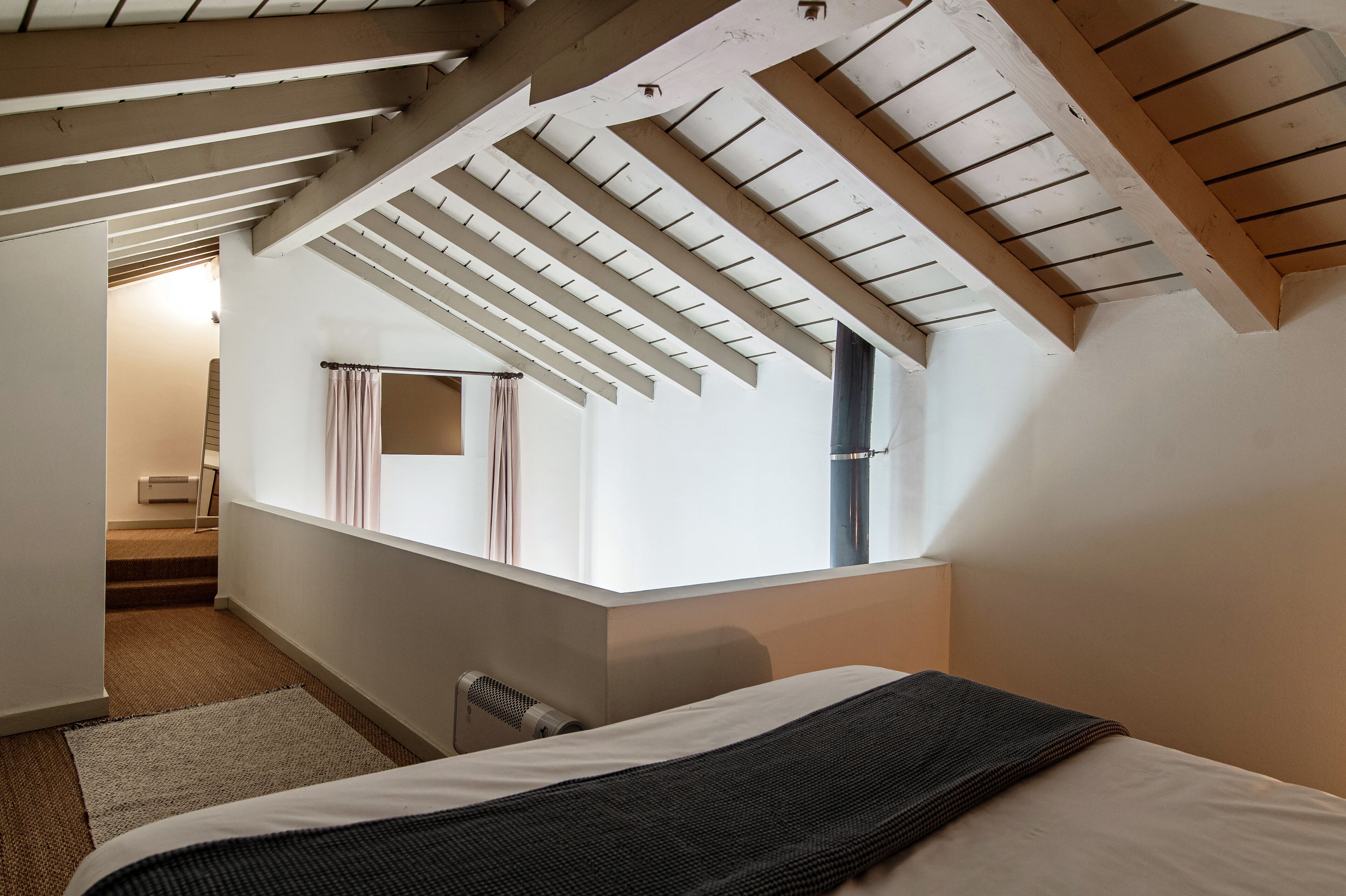 A bedroom with a sloped ceiling, a bed, a window with curtains, and a radiator.