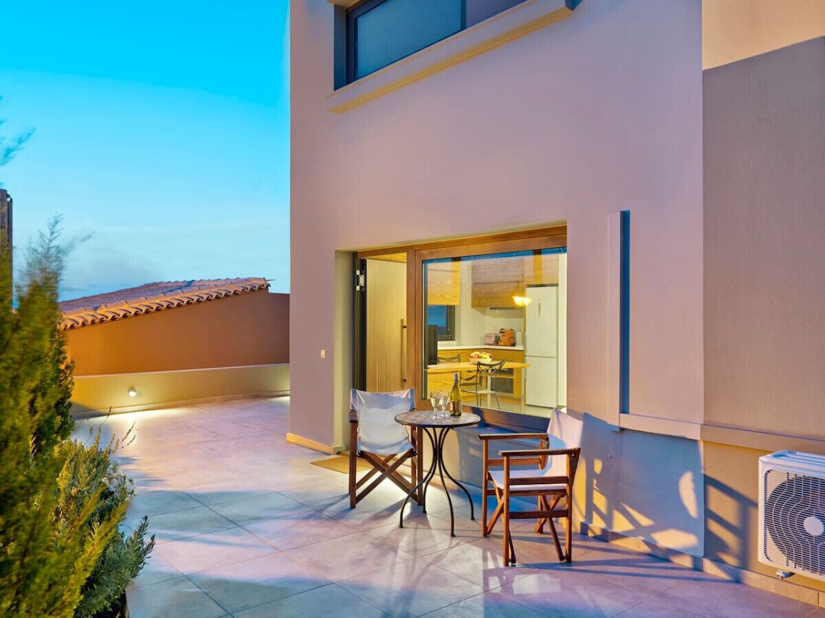 A modern building with a patio area featuring a table and chairs, a fan, and a view of the interior kitchen.