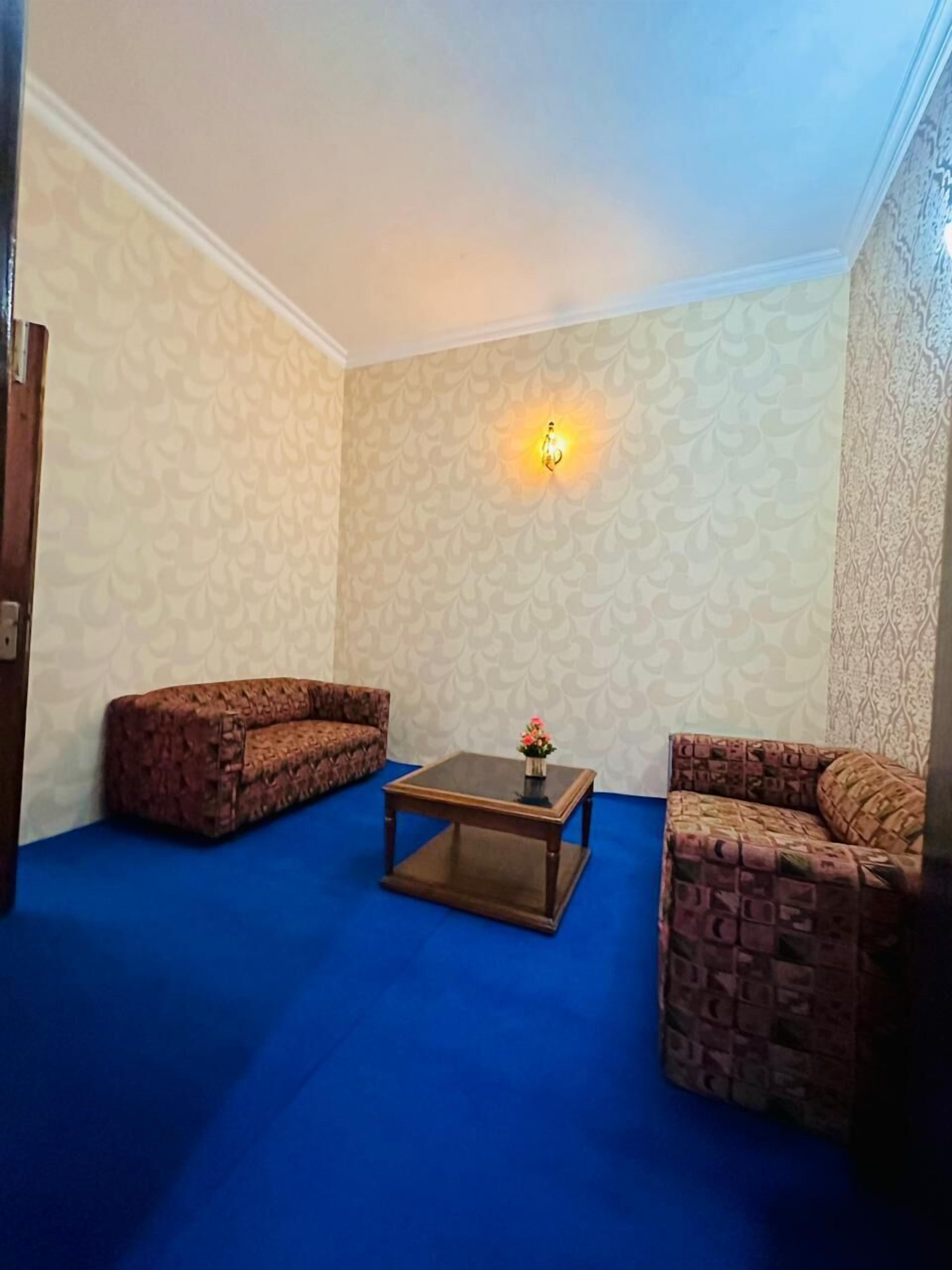 A room with patterned wallpaper, a blue carpet, two patterned sofas, a central coffee table with a flower vase, and a wall-mounted light fixture.