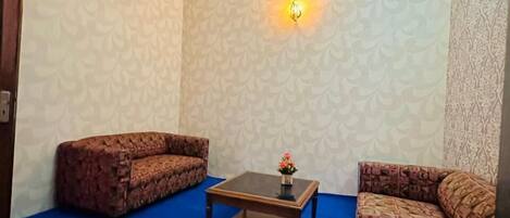 Una habitación con papel tapiz estampado, alfombra azul, dos sofás con diseños y una mesa de centro adornada con un jarrón de flores, además de una lámpara fijada a la pared.