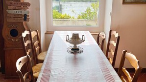 A dining table with a checkered tablecloth, a decorative teapot, and wooden chairs. A window with a view of a river and buildings is visible.