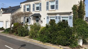 A two-story house with a dark roof, white walls, and blue shutters. There is a garden with various shrubs and a tree in front of the house.