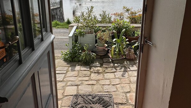A view from a room with a window overlooking a river, a stone pathway, and a variety of potted plants.