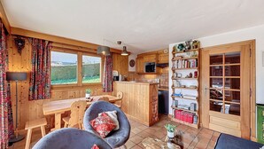 A cozy cabin-style interior with a wooden dining table, two chairs, a kitchen area with wooden cabinets, and a bookshelf filled with books and decorative items. - Charming 2BR Apartment with Mont Blanc Views, Terrace & Parking (Demi-Quartier)