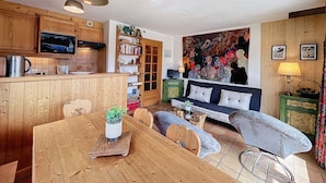 A cozy living room with a wooden sofa, a dining table with a plant, and a kitchen area with wooden cabinets and a TV. - Charming 2BR Apartment with Mont Blanc Views, Terrace & Parking (Demi-Quartier)