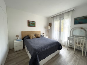A bedroom with a bed, a bedside table, a desk with a chair, a window with curtains, and a painting on the wall.