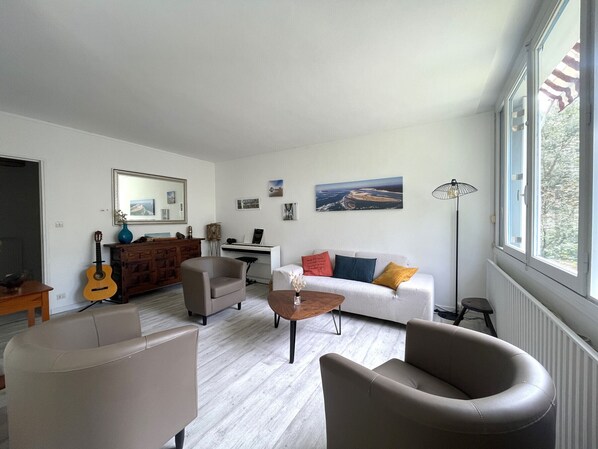 A living room with a white sofa, two armchairs, a coffee table, a guitar, a painting, and a radiator.