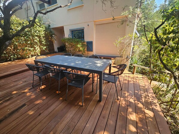 Una terraza de madera con una mesa y sillas, rodeada de follaje y un edificio con garaje.