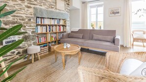 A cozy living room with a stone wall, a bookshelf, a grey sofa, a round wooden table, and wicker furniture.