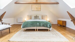 A bedroom with a sloped ceiling, wooden beams, a bed with a green blanket, two bedside tables with lamps, and a large window.