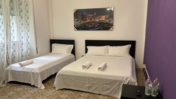 A hotel room with two single beds, a nightstand with a remote control and water bottles, and a cityscape painting on the wall. - VENICE NIGHT 25 FAMILY ROOM D (Mestre)