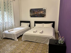 A hotel room with two single beds, a nightstand with a remote control and water bottles, and a cityscape painting on the wall. - VENICE NIGHT 25 FAMILY ROOM D (Mestre)