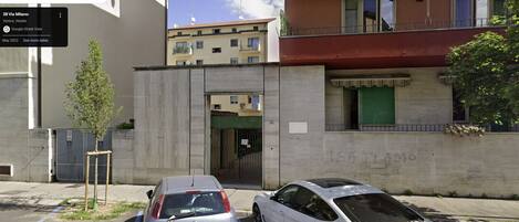 A street view with a building that has a green door and a sign with "IS ITAMO" written on the wall. There are two cars parked in front of the building.
