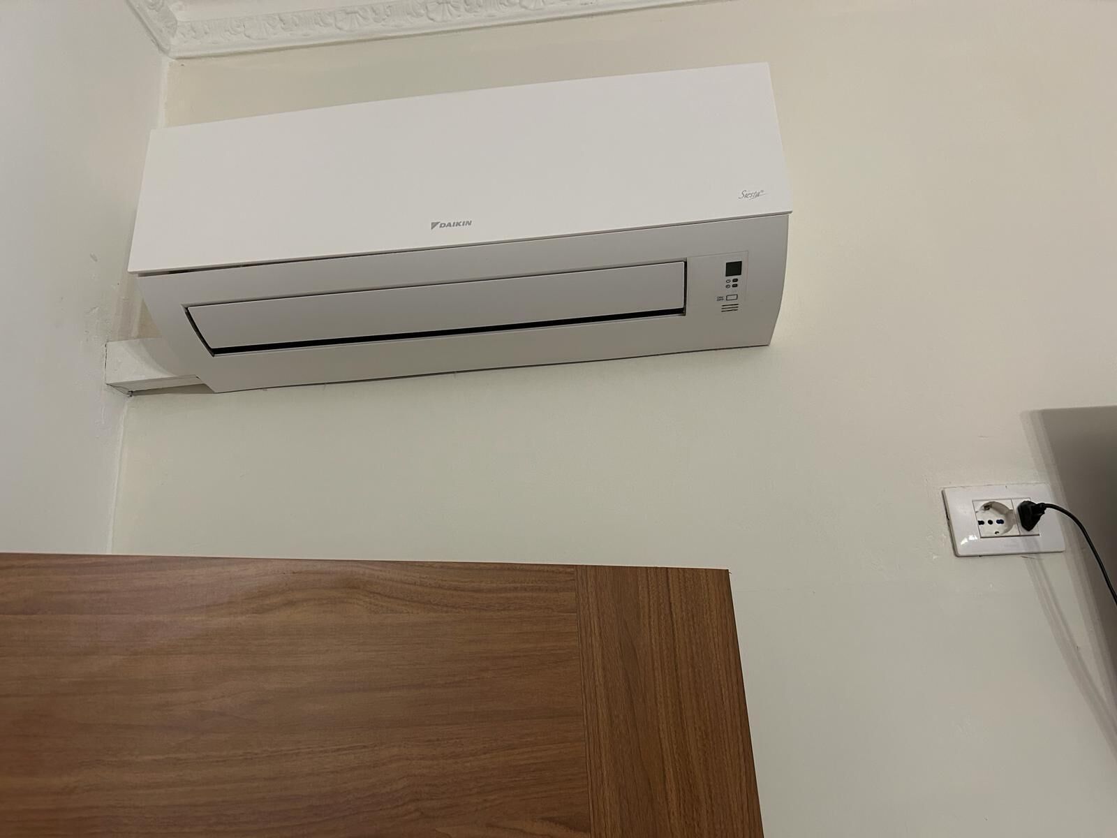 Aire acondicionado Daikin blanco montado en una pared blanca, con un enchufe a la derecha.