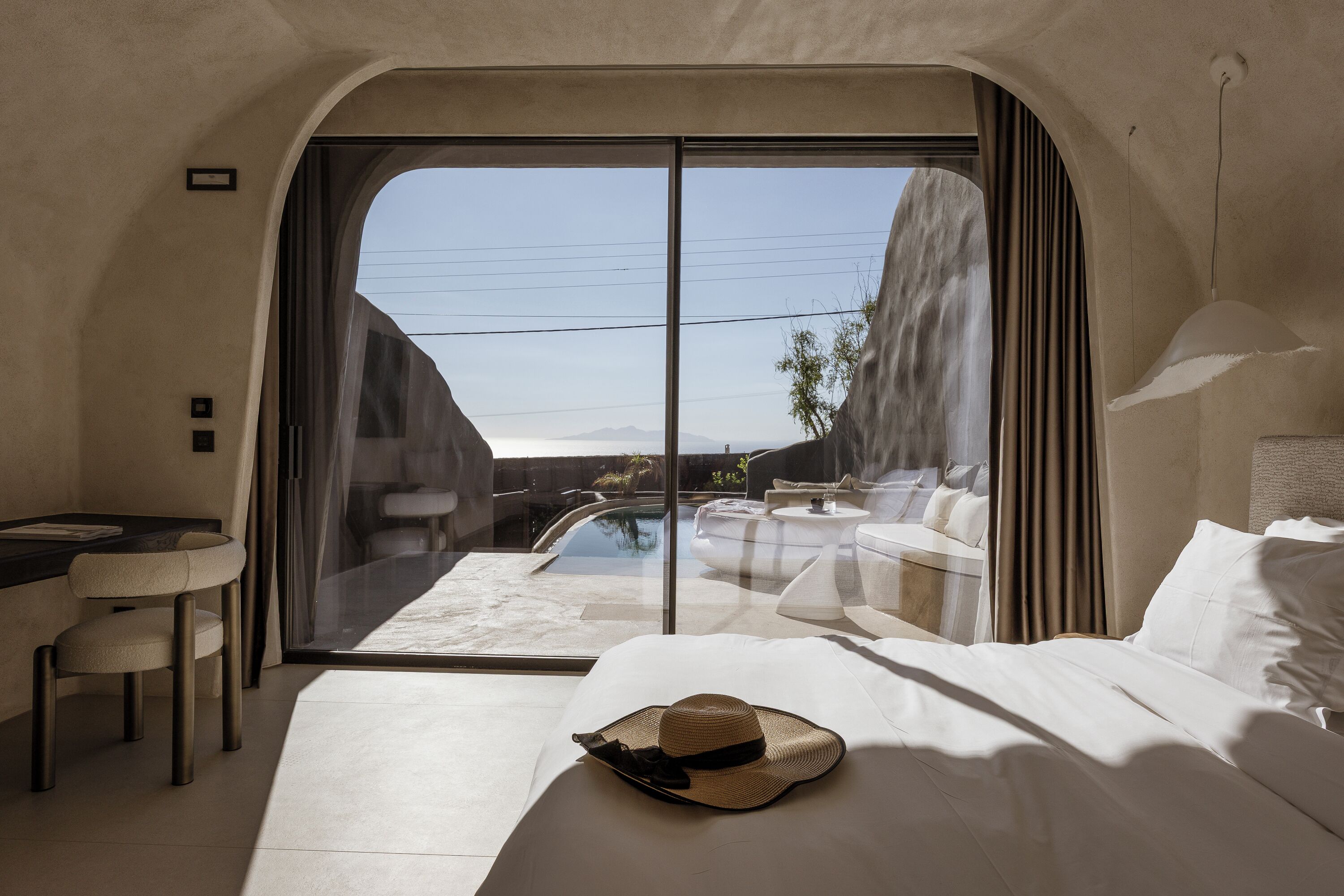 A modern hotel room with a large sliding glass door leading to a pool area, a bed with a hat on it, and a small table with a chair.