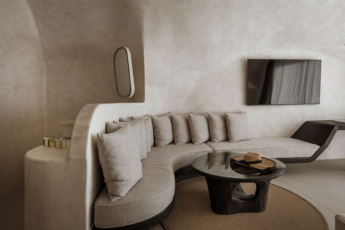 A modern living room with a curved sofa, a round coffee table, a wall-mounted TV, and a small side table with bottles.