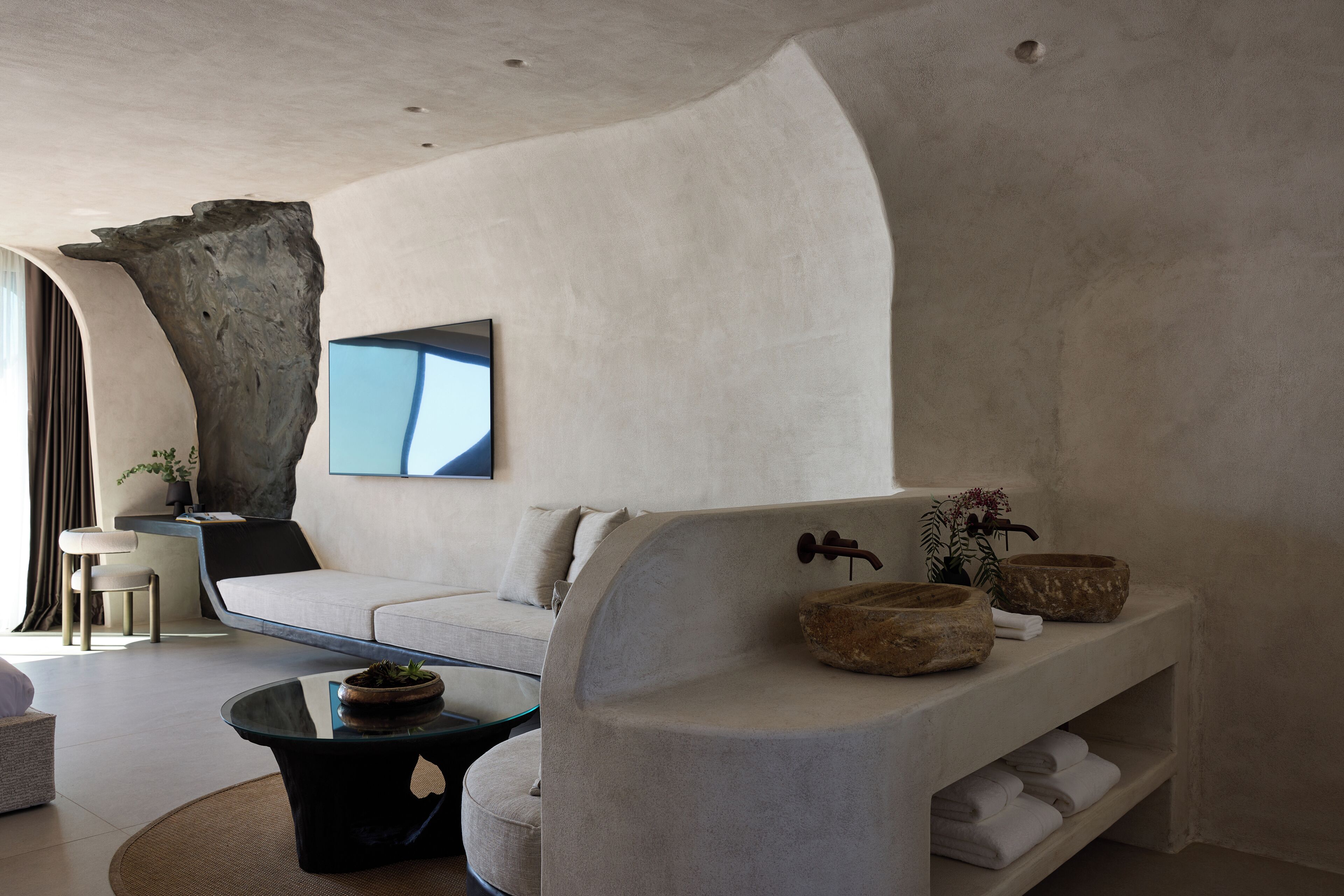 Un salón moderno con un sofá curvo, una mesa de centro de vidrio y una repisa de piedra con toallas y objetos decorativos.