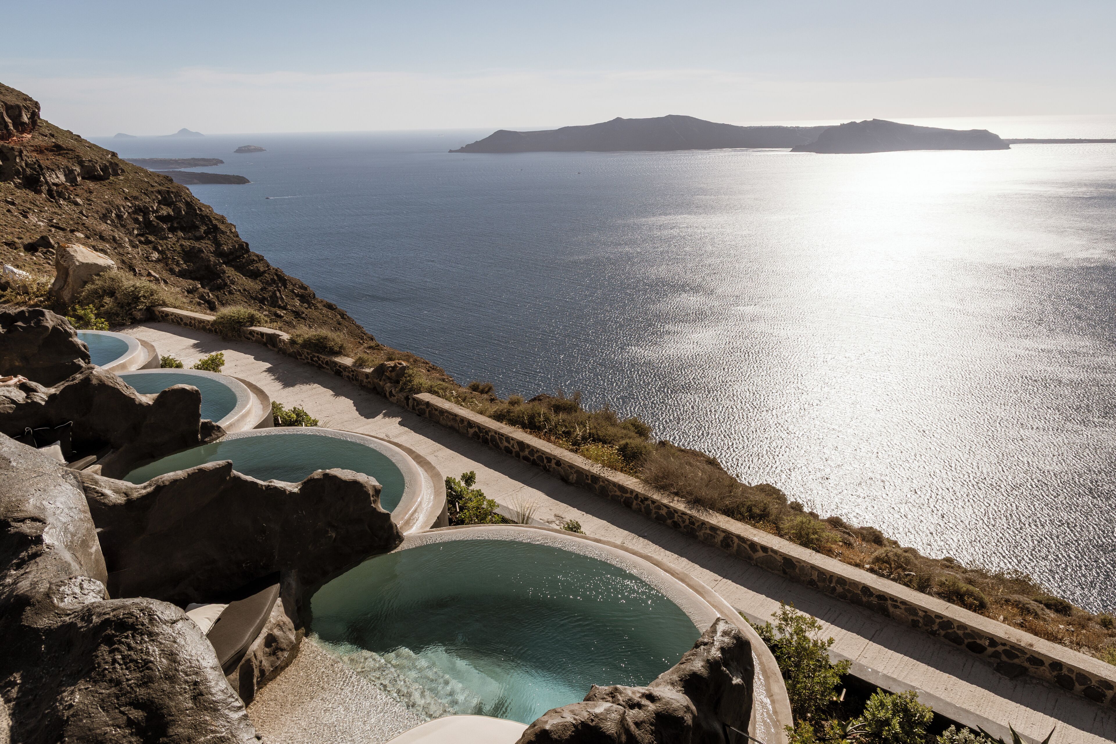 A coastal resort with multiple swimming pools, rocky terrain, and a view of the sea and distant islands.