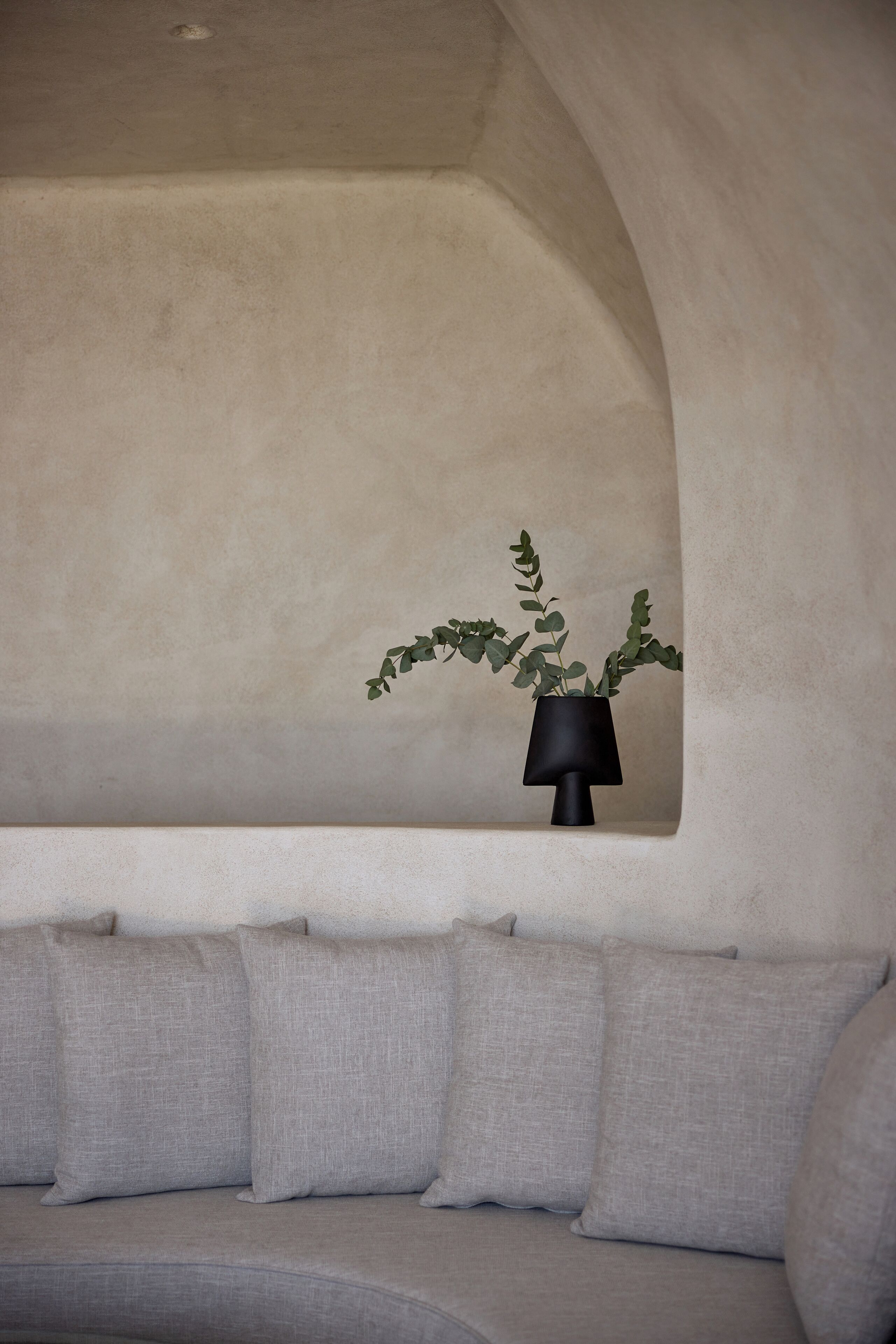 A modern interior with a curved ceiling, a black vase with green foliage, and a grey sofa with matching cushions.