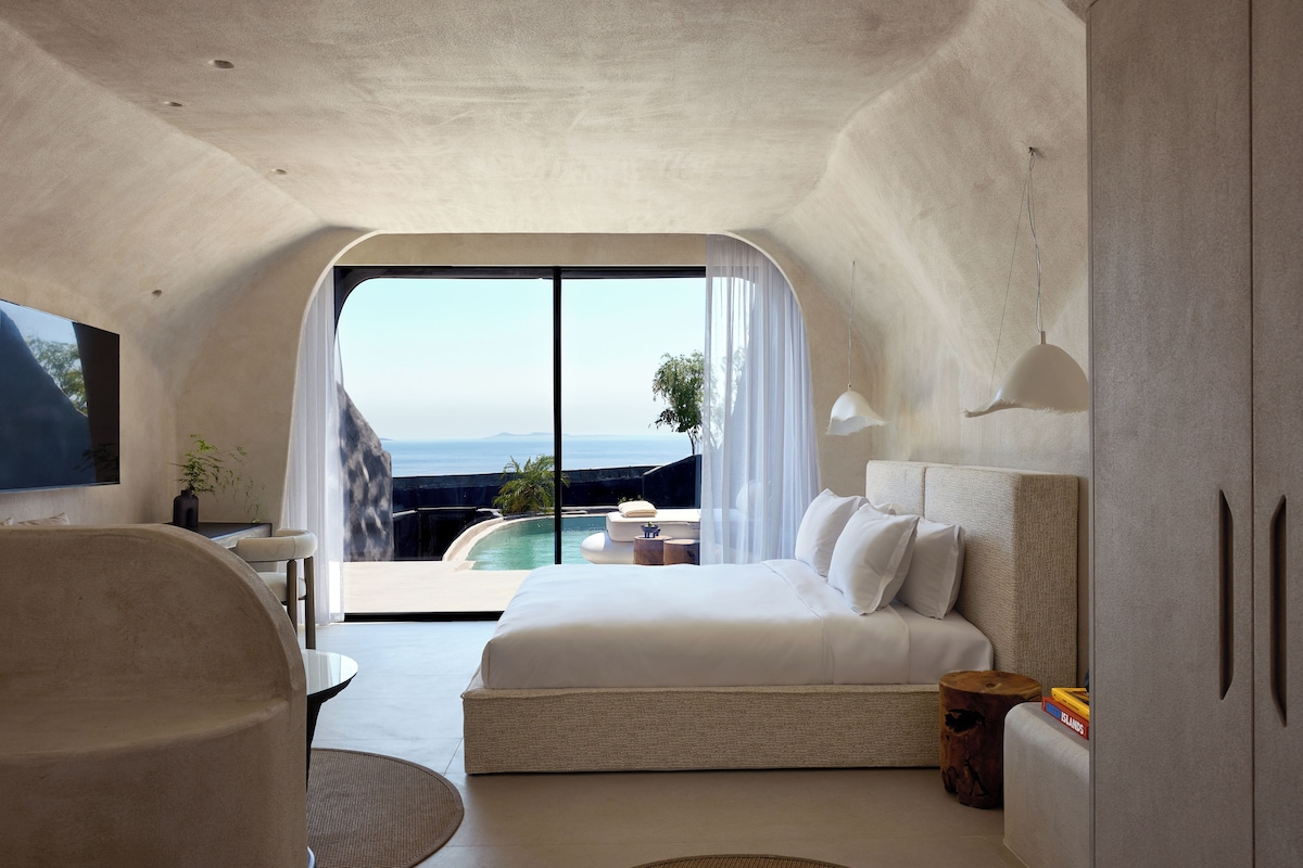 A modern bedroom with a large bed, a TV, a dining area, and a view of the ocean.