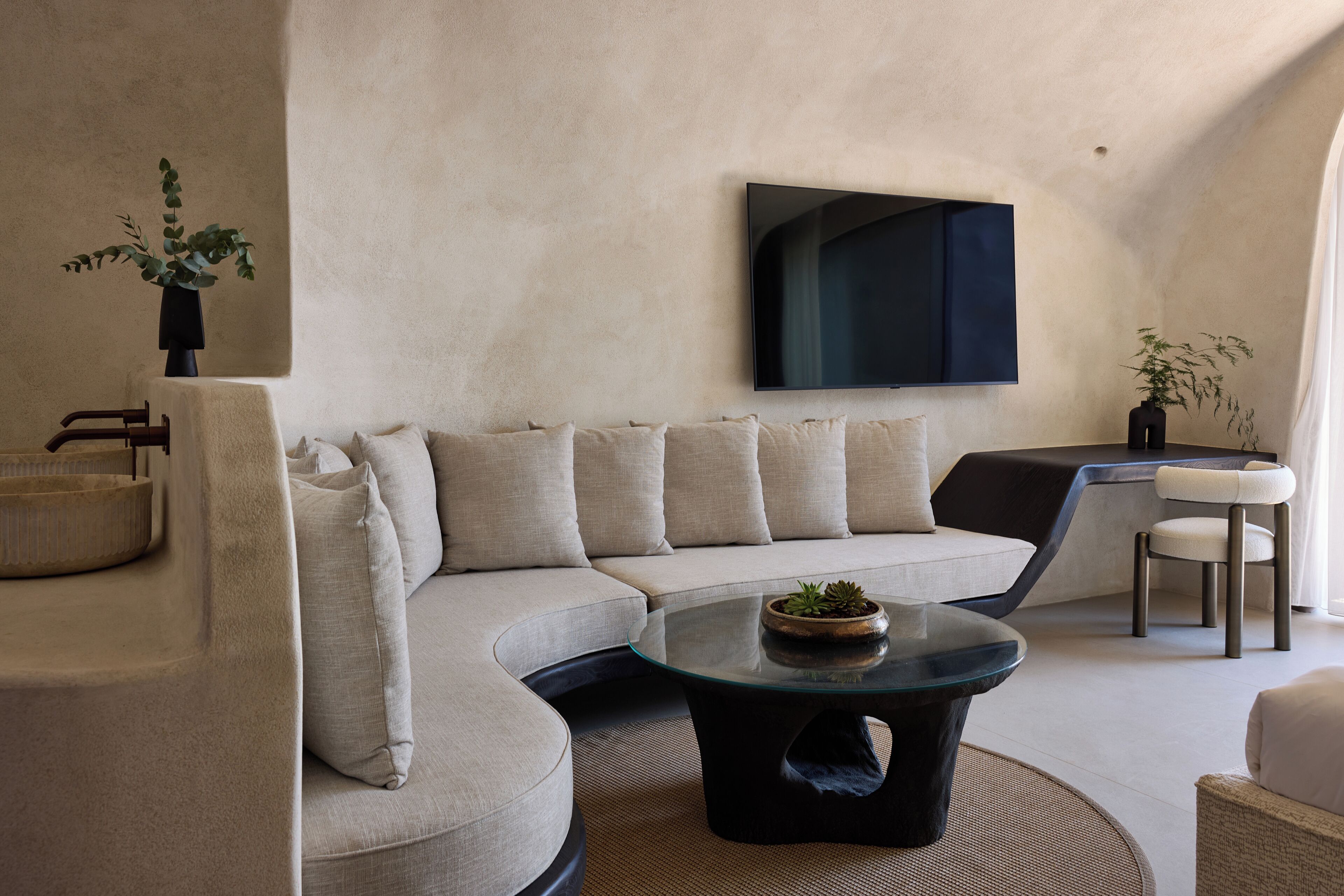 A modern living room with a curved sofa, a glass coffee table, and a flat-screen TV mounted on the wall.