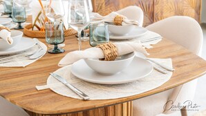 Una mesa de comedor dispuesta con platos, tazones y cubiertos blancos, servilletas tejidas y cristalería transparente.