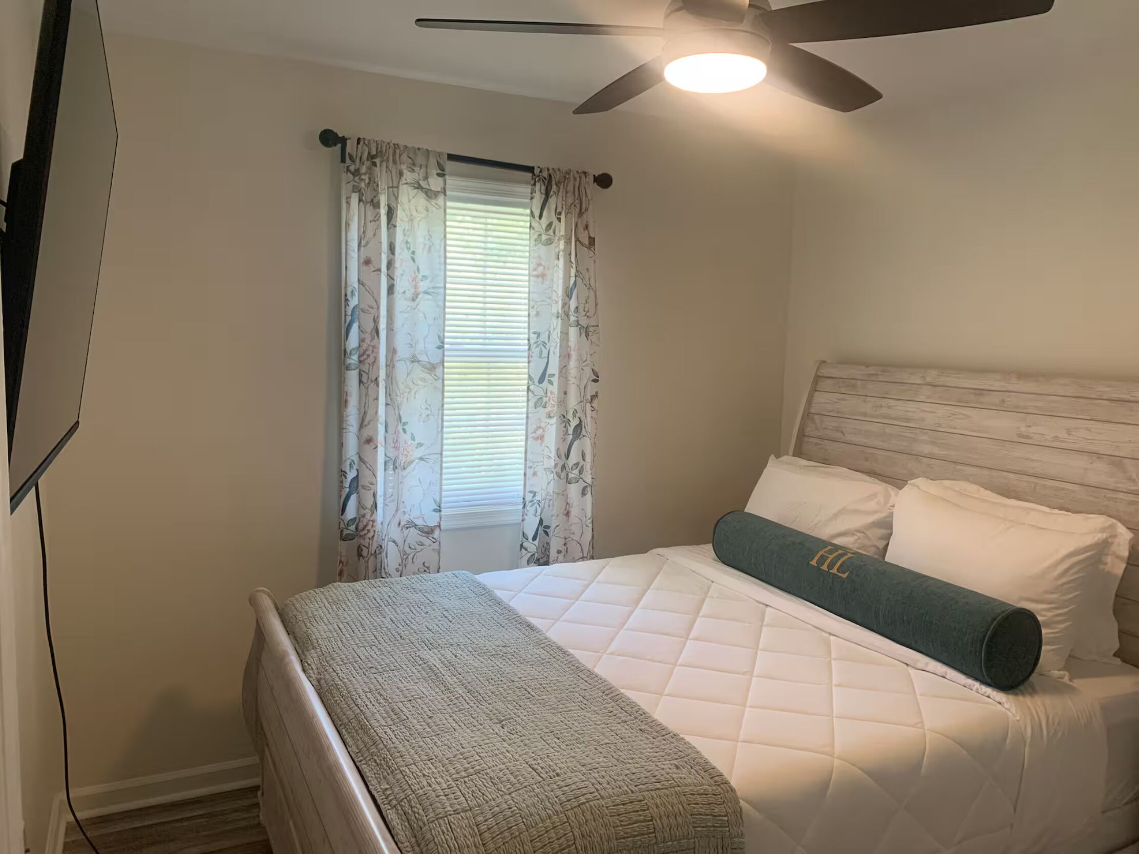 A bedroom with a bed, a ceiling fan, and a window with floral curtains.