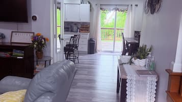 Een moderne woonkamer met een grijze bank, een eettafel met een kant kleedje, en een keuken op de achtergrond.