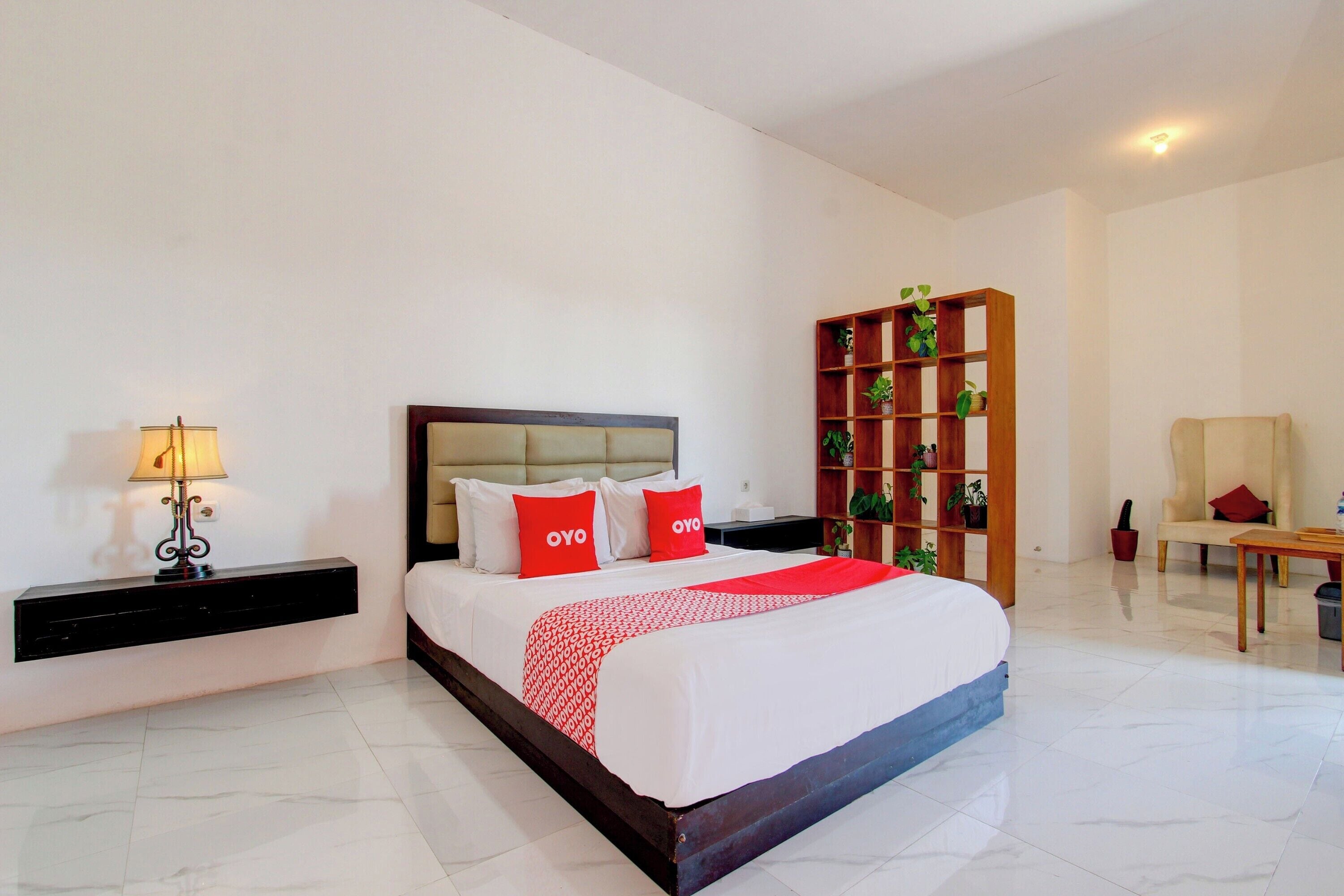 Una habitación de hotel con una cama grande, una mesita de noche con lámpara, una estantería de madera y una silla.