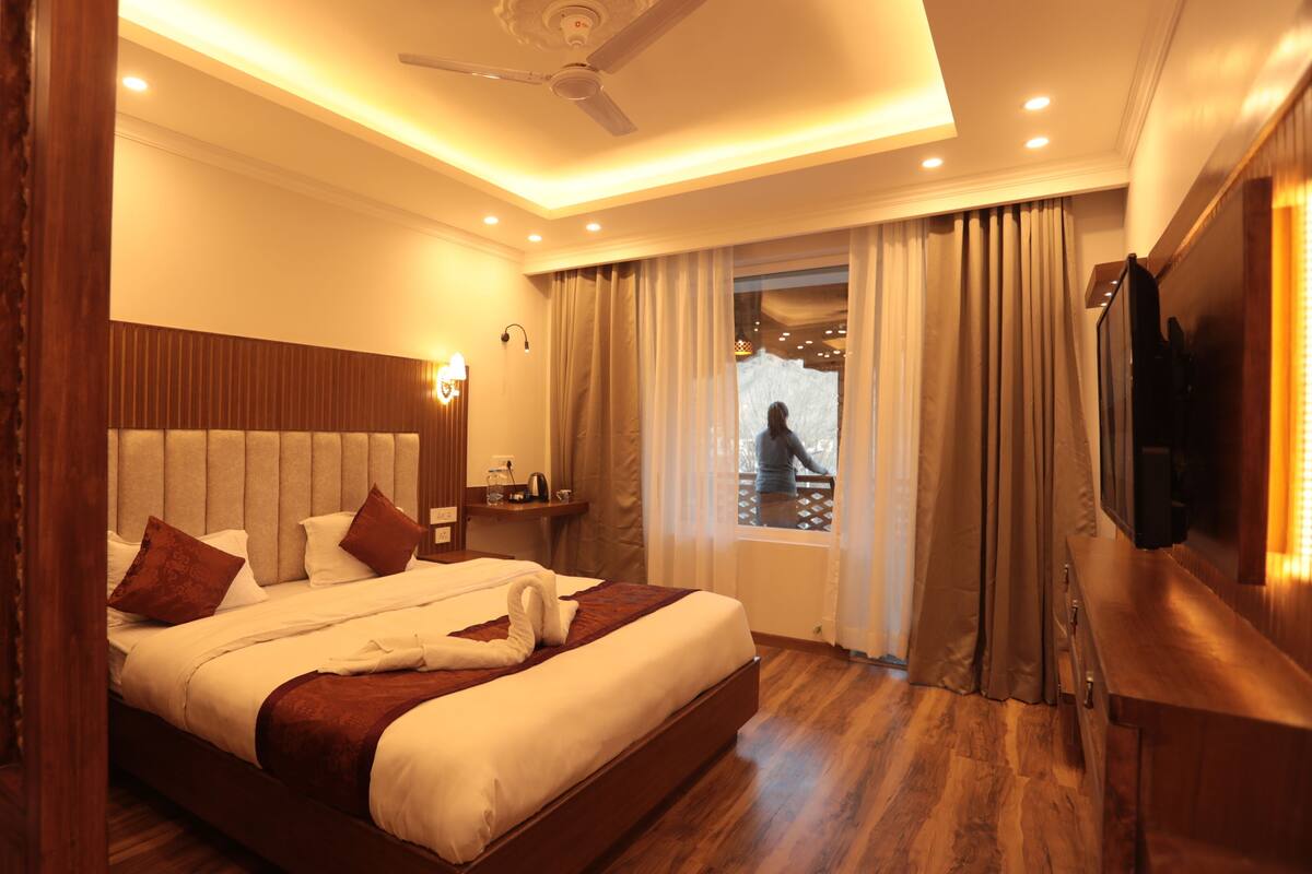 Una habitación de hotel con una cama grande, cabecera de madera, una mesita de noche con lámpara, televisión y vistas a un balcón.