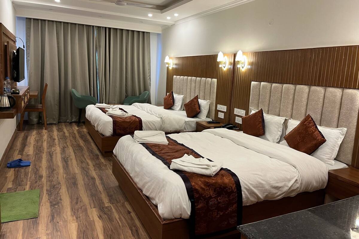 Habitación de hotel con tres camas, un escritorio con silla, televisor y un ventanal con cortinas.
