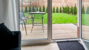 A modern room with a sliding glass door leading to a patio with a table and chairs, a brick wall, and a grassy area with trees. - Hideaway Apartment - Terrace with Garden & Parking by Rentujemy (Lódz)