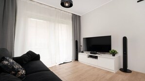 A modern living room with a black sofa, a flat-screen TV on a white console, and a large window with sheer curtains. - Hideaway Apartment - Terrace with Garden & Parking by Rentujemy (Lódz)