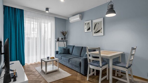 A modern living room with a blue sofa, a dining table with chairs, a TV, and a large window with curtains. - Warsaw Airport Studio with Private Terrace by Rentujemy (Warszawa)