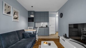 A modern living room with a dark grey sofa, a white coffee table with pastries, a dining area with a white table and chairs, a kitchen with grey cabinets, and a flat-screen TV on a white stand. - Warsaw Airport Studio with Private Terrace by Rentujemy (Warszawa)