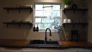 A kitchen with a window, a sink, and shelves with dishes and bottles. - Peaceful Cabin Steps Away From The Creek, Convenient Access To ATV Trails! (Mena)